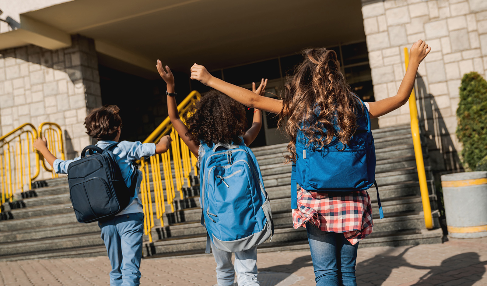 Three kids with backpacks raise their arms as they head up the steps to school.